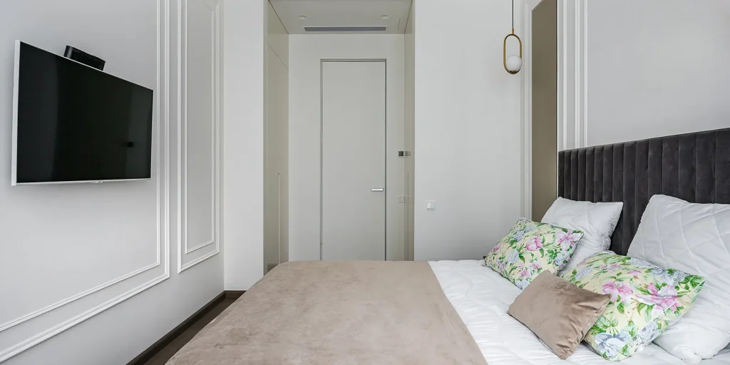 A large comfortable double bed with pillows, with a TV opposite and a half-mounted lamp in a small white and bright bedroom in a modern apartment